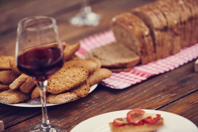 Verre de vin rouge accompagné de crackers dorés, pain artisanal tranché et bruschetta tomate sur une table en bois rustique.