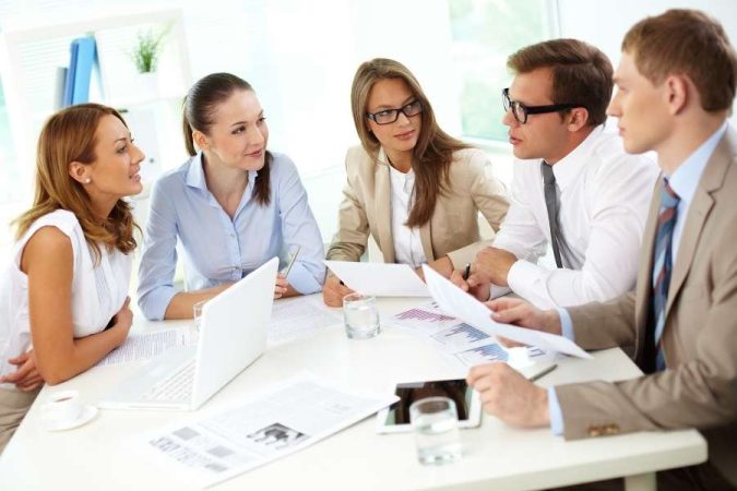 Un groupe de cinq professionnels en tenue d'affaires discute activement autour d'une table de réunion blanche.